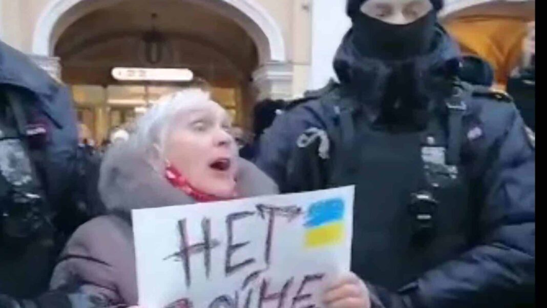 'n bejaarde vrou met wit hare hou 'n plakkaat vas teen die oorlog in die Oekraïne wat deur twee Russiese polisiemanne gearresteer is